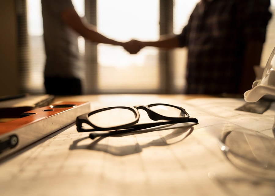 Two business professionals shaking hands over a table with eyewear designs and material samples.